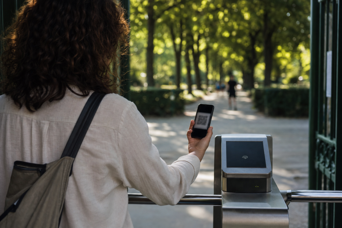 Besucherin hält Smartphone mit QR-Code-Ticket am Eingang eines Berliner Parks, grüne Baumallee und Eingangstor im Hintergrund