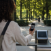 Besucherin hält Smartphone mit QR-Code-Ticket am Eingang eines Berliner Parks, grüne Baumallee und Eingangstor im Hintergrund